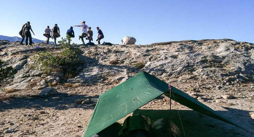 A group of outward bound students stand in a circle and stretch. There is a tarp shelter in the foreground.